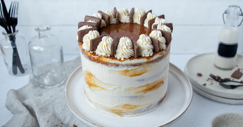 Hohe, elegant verzierte Geburtstagstorte im Naked-Cake-Stil mit heller Creme und Karamell. Oben auf der Torte befindet sich ein glänzender Karamellspiegel, umrandet von Sahnetuffs und Daim Minis. Die Torte steht auf einem schlichten, weißen Keramikteller. Im Hintergrund Glasflaschen, Besteck und verstreute Schokostückchen auf einem hellen Tisch.