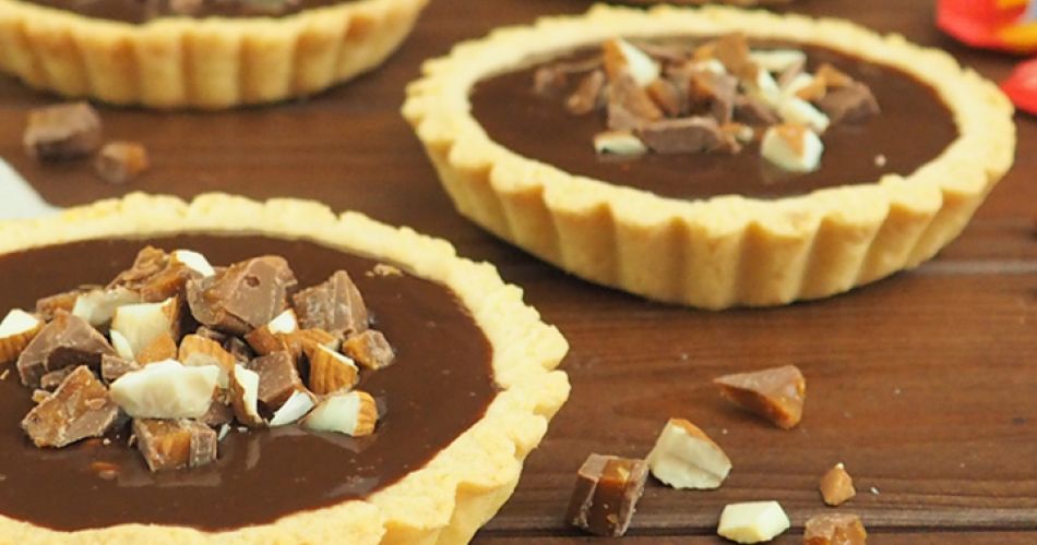 Vier kleine Mürbeteig-Tartelettes mit glänzender Daim Schokoladenfüllung stehen auf einer Holzoberfläche. Jedes Tartelette ist mit grob gehackten Daim-Stückchen und Mandeln bestreut. Im Vordergrund liegt ein beiger Stoff, auf dem sich ebenfalls kleine Schokoladenstücke befinden.