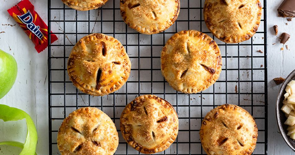 Frisch gebackene, runde Mini-Apple-Pies mit goldbrauner Kruste liegen auf einem schwarzen Abkühlgitter. Einige der Pies haben karamellisierte Daim-Stückchen auf der Oberfläche. Links daneben grüne Apfelschalen und ein Messer, oben und unten mehrere verpackte Daim-Riegel. In einer Holzschale rechts oben liegen ungeschmolzene Daim-Stückchen.