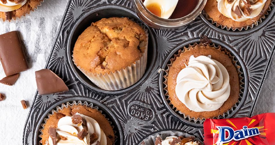 Sechs Muffinmulden einer dekorativen Backform, fünf sind mit verschiedenen Daim-Cupcakes gefüllt. Einige sind mit Sahnecreme, Karamell und Daim-Stückchen verziert, einer ist halbiert und zeigt eine Karamellfüllung. In einer Mulde liegen Daim-Riegel, auf der Backform steht noch ein Glas mit Karamellsauce und Löffel. Außen herum verstreute Daim-Riegel und Schokoladenstücke.