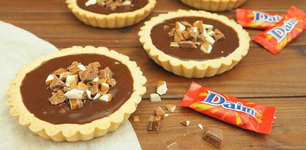 Drei Daim-Tartelettes mit Daim Schokoladenfüllung und gehackten Daim-Stückchen und Mandeln stehen auf einer Holzfläche. Im Vordergrund liegen rote verpackte Daim-Minis und verstreute Schoko- und Karamellstücke. Links unten liegt ein Tartelette auf einem Leinentuch