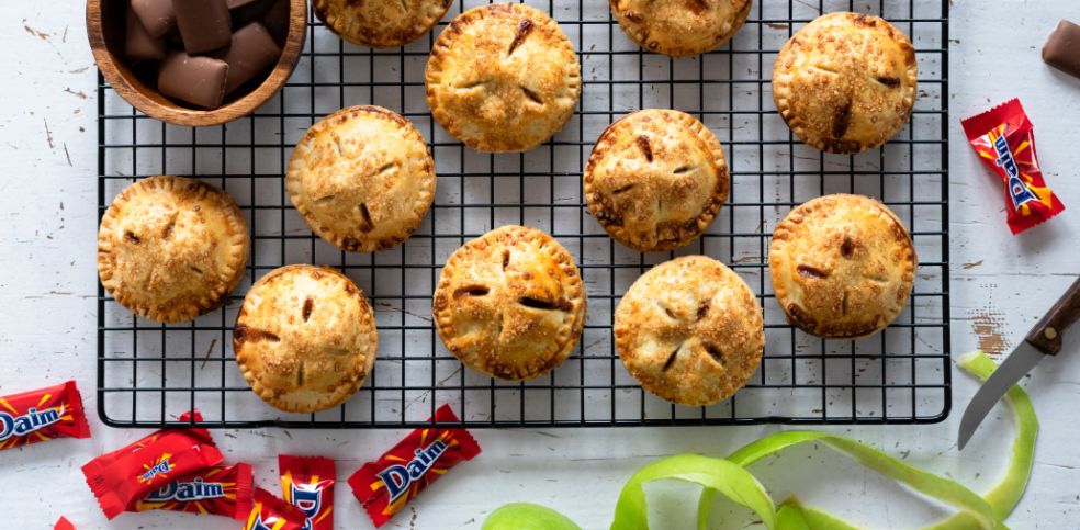 Frisch gebackene, runde Mini-Apple-Pies mit goldbrauner Kruste liegen auf einem schwarzen Abkühlgitter. Einige der Pies haben karamellisierte Daim-Stückchen auf der Oberfläche. Links daneben grüne Apfelschalen und ein Messer, oben und unten mehrere verpackte Daim-Riegel. In einer Holzschale rechts oben liegen ungeschmolzene Daim-Stückchen.