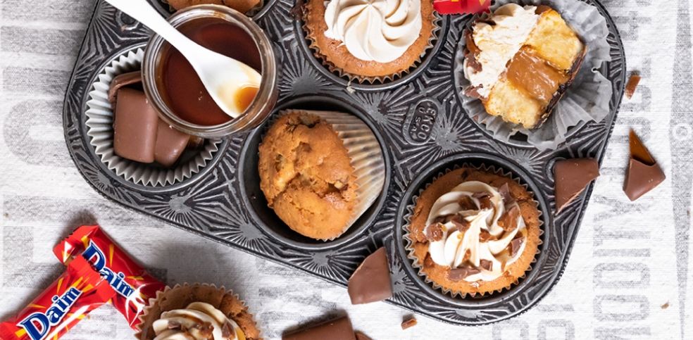 Sechs Muffinmulden einer dekorativen Backform, fünf sind mit verschiedenen Daim-Cupcakes gefüllt. Einige sind mit Sahnecreme, Karamell und Daim-Stückchen verziert, einer ist halbiert und zeigt eine Karamellfüllung. In einer Mulde liegen Daim-Riegel, auf der Backform steht noch ein Glas mit Karamellsauce und Löffel. Außen herum verstreute Daim-Riegel und Schokoladenstücke.