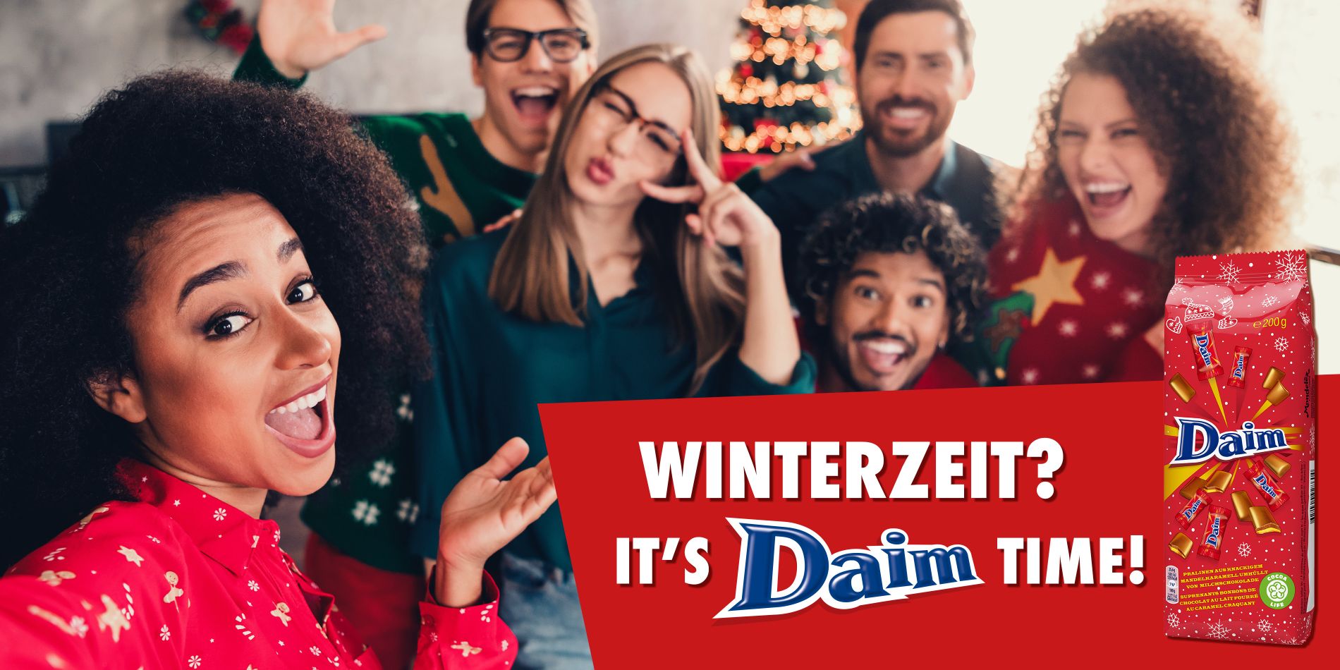 Eine Gruppe fröhlicher Menschen in weihnachtlichen Pullovern posiert für ein Selfie; im Vordergrund hält eine Person die Kamera. Rechts im Bild befindet sich ein Werbebanner mit der Aufschrift ‚Winterzeit? It’s Daim Time!‘ sowie einer roten Daim-Verpackung mit weihnachtlichem Design.
