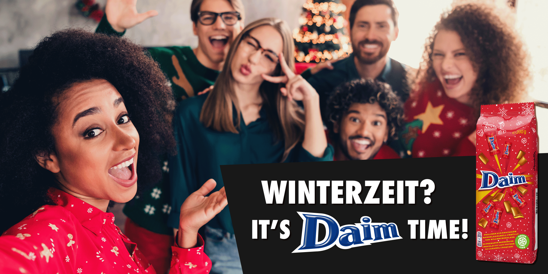 Eine Gruppe fröhlicher Menschen in weihnachtlichen Pullovern posiert für ein Selfie; im Vordergrund hält eine Person die Kamera. Rechts im Bild befindet sich ein Werbebanner mit der Aufschrift ‚Winterzeit? It’s Daim Time!‘ sowie einer roten Daim-Verpackung mit weihnachtlichem Design.